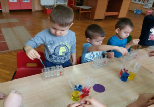 dzieci siedzą przy stolikach i pipetami nabierają kolory i w lewaja do probówek